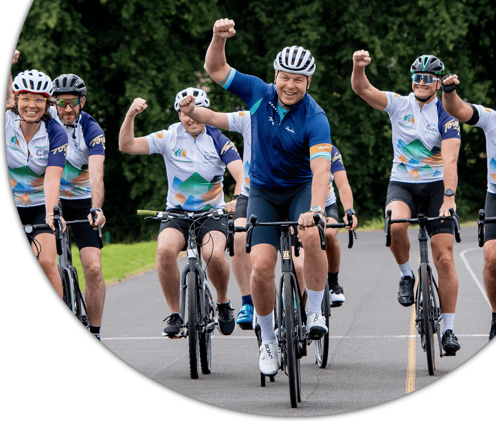 A group of cyclists in matching jerseys and helmets ride along a paved road, smiling and raising their fists in celebration during a charity bike ride, with Sir Chris Hoy at the centre front of the group.